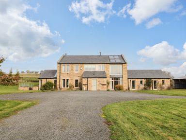 A stone farmhouse with a gravel driveway and green lawn under a blue sky at Fontburn Farmhouse in Ewesley near Rothbury