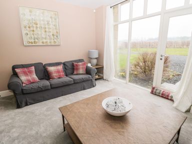 A living room with a gray sofa and plaid cushions a wooden coffee table and large windows at Fontburn Farmhouse in Ewesley near Rothbury