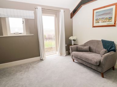 A small seating area with a gray sofa pillows and a throw blanket next to a window with white curtains and a side table with flowers at Fontburn Farmhouse Ewesley near Rothbury