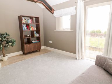 A room with a wooden bookshelf filled with books a potted plant a window with a blind a glass door with curtains and a grey sofa at Fontburn Farmhouse in Ewesley near Rothbury