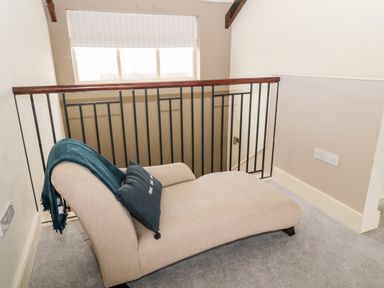 A beige chaise lounge with a dark pillow and blanket near a black metal railing and window in a carpeted room at Fontburn Farmhouse in Ewesley near Rothbury
