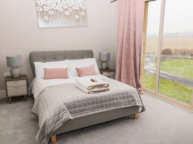 A bedroom with a grey bed a quilt pink pillows bedside tables with lamps and large window with pink curtains at Fontburn Farmhouse in Ewesley near Rothbury