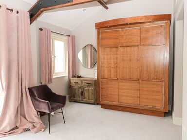 A room with a large wooden wardrobe a wooden cabinet a unique wall mirror a dark upholstered chair and pink curtains at Fontburn Farmhouse Ewesley near Rothbury