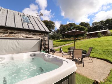 An outdoor patio with a hot tub a table with four chairs and an umbrella next to a stone building with a metal roof at Y Felin at Coed Cadw in Felindre Farchog near Newport Pembrokeshire