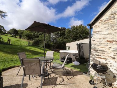 An outdoor patio with a table and four chairs under an umbrella a barbecue grill and a hot tub next to a stone building at Y Felin at Coed Cadw in Felindre Farchog near Newport Pembrokeshire