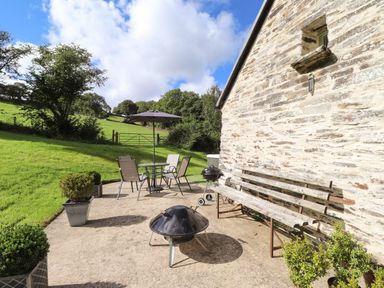 An outdoor patio with a metal table and chairs with an umbrella a barbecue grill several potted plants and a wooden bench by a stone wall at Y Felin at Coed Cadw in Felindre Farchog near Newport Pembrokeshire