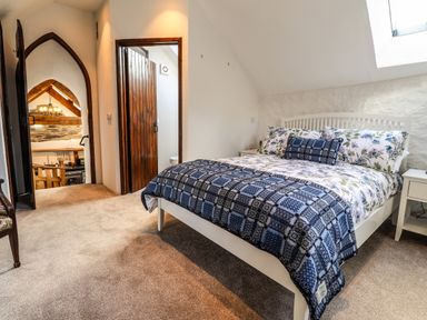 A bedroom with a double bed featuring floral and checkered bedding two white bedside tables with lamps a wooden door leading to a bathroom and a doorway showing a kitchen area at Y Felin at Coed Cadw in Felindre Farchog near Newport Pembrokeshire