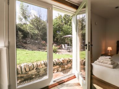 A bedroom with a bed and folded towels near an open glass door leading to a garden with a stone wall and outdoor seating at Y Felin at Coed Cadw in Felindre Farchog near Newport Pembrokeshire