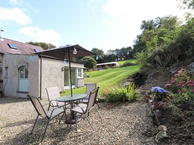 A gravel patio with a glass table and four chairs under an umbrella beside a stone house with a garden and grassy hill in the background at Y Felin at Coed Cadw in Felindre Farchog near Newport Pembrokeshire