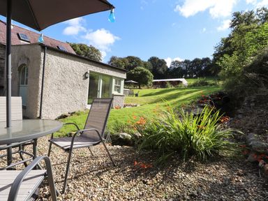 An outdoor patio with chairs and a table next to a stone house and a grass lawn with trees at Y Felin at Coed Cadw in Felindre Farchog near Newport Pembrokeshire