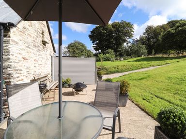 An outdoor patio with a glass table, umbrella, chairs, fire pits, and a stone wall next to a grassy area with trees at Y Felin at Coed Cadw in Felindre Farchog near Newport Pembrokeshire