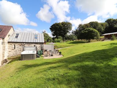 A backyard with a patio table and chairs an outdoor hot tub trees and a stone building at Y Felin at Coed Cadw in Felindre Farchog near Newport Pembrokeshire