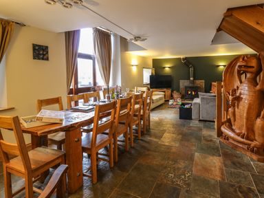A dining area with a wooden table and chairs and a living area with sofas a wood stove and a carved wooden dragon at Maes Y Berllan Barn in Gilwern