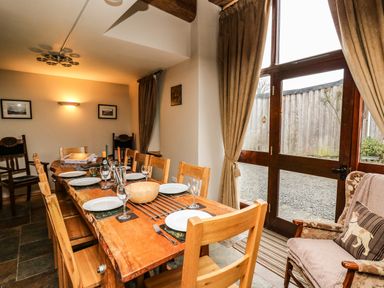 A dining room with a wooden table set with plates and glasses near large windows with curtains at Maes Y Berllan Barn in Gilwern