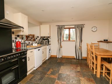 A kitchen with a dining table set wooden chairs a window with striped curtains and various kitchen appliances at Maes Y Berllan Barn in Gilwern