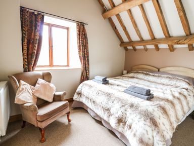 A bedroom with a double bed covered with a fur blanket two folded towels on the bed a cushioned armchair and a window with patterned curtains at Maes Y Berllan Barn in Gilwern