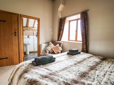 A bedroom with a bed covered in a fur blanket and folded towels two cushions on a chair near a window and a bathroom visible through an open door at Maes Y Berllan Barn in Gilwern