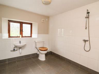 A bathroom with a toilet with wooden seat a wall-mounted sink a shower head on tiled wall and a window at Maes Y Berllan Barn in Gilwern
