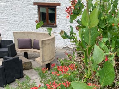 An outdoor patio with woven chairs a curved stone bench with cushions and flowering plants at Maes Y Berllan Barn in Gilwern