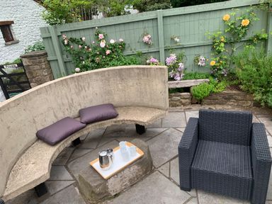 An outdoor seating area with a curved stone bench with cushions a wicker armchair a tray with a teapot and cups on a stone table and a green wooden fence with flowers at Maes Y Berllan Barn in Gilwern
