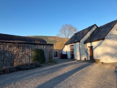 An exterior view of a white stone building with a gravel driveway and a wooden barn with hills in the background at Maes Y Berllan Barn in Gilwern