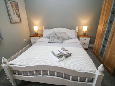 A bedroom with a white bed with pillows and folded towels on top two bedside tables with lamps a wardrobe and a painting on the wall at Pebble Lodge in Kenmare County Kerry