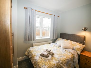 A bedroom with a double bed with folded towels on top a wooden wardrobe window with curtains and a bedside table with a lamp at Pebble Lodge in Kenmare County Kerry