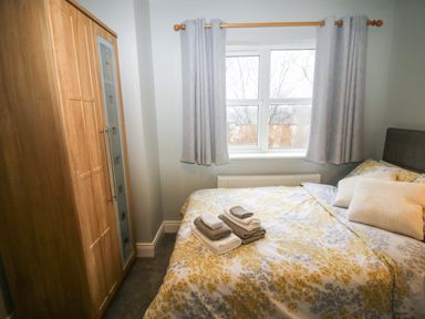 A bedroom with a wooden wardrobe a bed with patterned bedding and folded towels near a window with curtains at Pebble Lodge Kenmare County Kerry