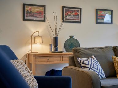 A living room with a wooden console table with lamp and vases and framed pictures on the wall with a blue armchair and a gray sofa with decorative cushions at Riverside Park 2 in Keswick