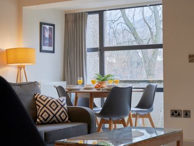 A dining area with a round table set with glasses of orange juice and bowls next to a living room with a sofa and a lamp at Riverside Park 2 in Keswick