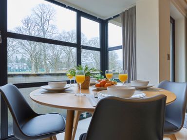 A dining table set with bowls glasses of orange juice and a fruit plate next to large windows with trees outside at Riverside Park 2 in Keswick