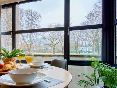 A dining table set with bowls croissants and glasses of orange juice beside large windows overlooking a park at Riverside Park 2 in Keswick