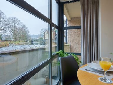 A corner dining area with a glass of orange juice on a wooden table near large windows overlooking a river and buildings at Riverside Park 2 in Keswick