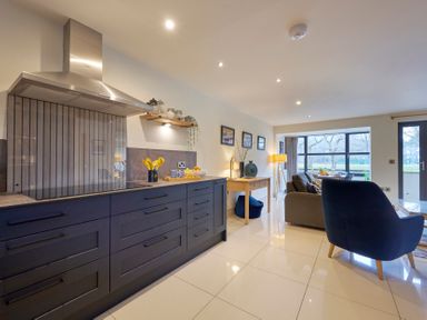 A kitchen area with dark cabinetry and a stainless steel range hood next to a living space with chairs a sofa and dining table by a large window at Riverside Park 2 in Keswick