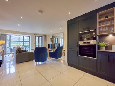 An open plan living room and kitchen with blue armchairs a grey sofa a dining table by windows and built in ovens at Riverside Park 2 in Keswick