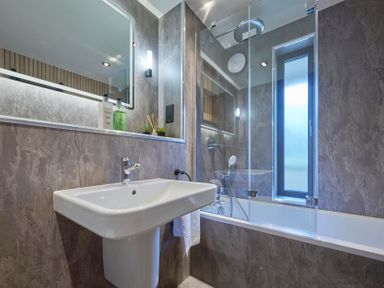 A bathroom with a white sink a bathtub with glass screen and a tall narrow window at Riverside Park 2 in Keswick