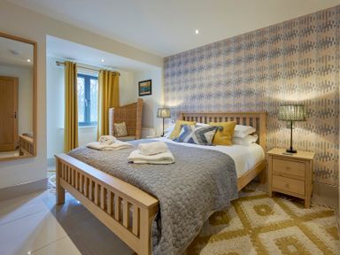 A bedroom with a wooden bed frame grey bedding yellow curtains bedside tables and a wicker chair at Riverside Park 2 in Keswick