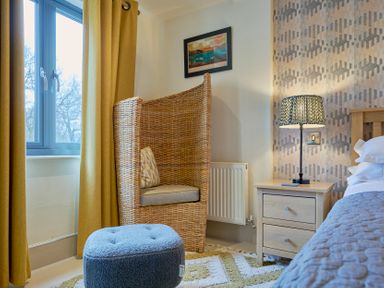 A bedroom corner with a wicker armchair a blue ottoman and a wooden bedside table with a lamp at Riverside Park 2 in Keswick