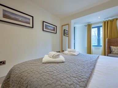 A bedroom with a bed covered with a gray quilt and folded white towels on top next to a window with yellow curtains and a wicker chair at Riverside Park 2 Keswick