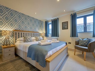A bedroom with a bed blue and white bedding two bedside tables with lamps an armchair and two windows at Riverside Park 2 in Keswick