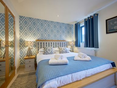 A bedroom with a double bed blue quilt and towels bedside tables with lamps and a window with blue curtains at Riverside Park 2 in Keswick