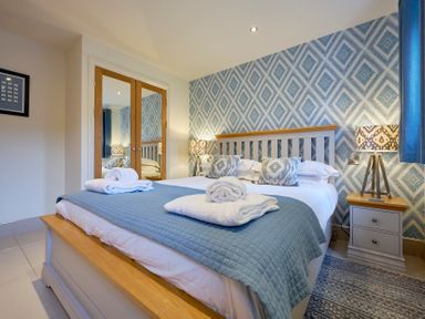 A bedroom with a double bed two bedside tables with lamps wardrobe with mirrored doors and patterned wallpaper at Riverside Park 2 Keswick