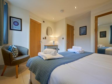 A bedroom with a bed covered in white sheets and a blue quilt with folded towels on top a small armchair with a pillow a bedside table with a lamp a mirror and two wooden doors at Riverside Park 2 in Keswick