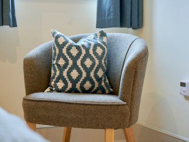 A grey upholstered chair with wooden legs and a patterned cushion in a room with blue curtains and a thermostat on the wall at Riverside Park 2 in Keswick