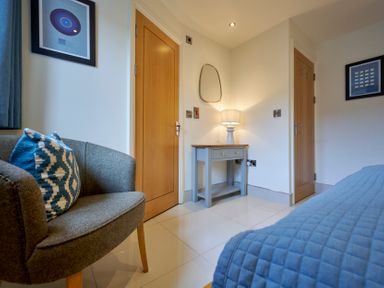 A bedroom corner with a gray armchair with a patterned cushion a blue quilted bedspread a small table with drawers and a lamp wooden doors and framed pictures on white walls at Riverside Park 2 Keswick