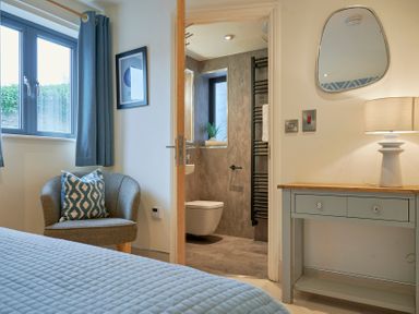 A bedroom with a grey chair next to a window and blue curtains a small desk with a lamp and an open door leading to a bathroom at Riverside Park 2 in Keswick