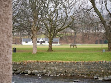A river with rocks and a stone wall alongside a grassy park with trees benches and a pavilion building at Riverside Park 2 in Keswick