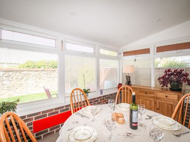 A dining area with a round table set with plates glasses and a wine bottle near windows with blinds and a wooden sideboard at Knapp Cottage in Chickerell