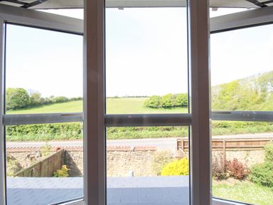 A view of green fields and trees through a window with a stone wall and road outside at Knapp Cottage in Chickerell