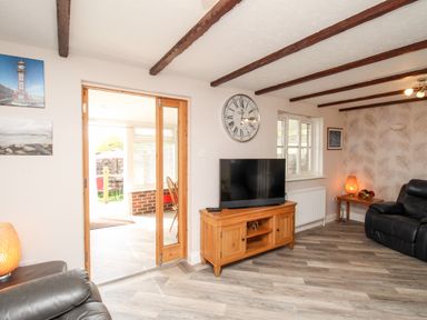 A living room with a wooden TV stand holding a television a large wall clock leather armchairs and a wooden side table with a lamp at Knapp Cottage in Chickerell